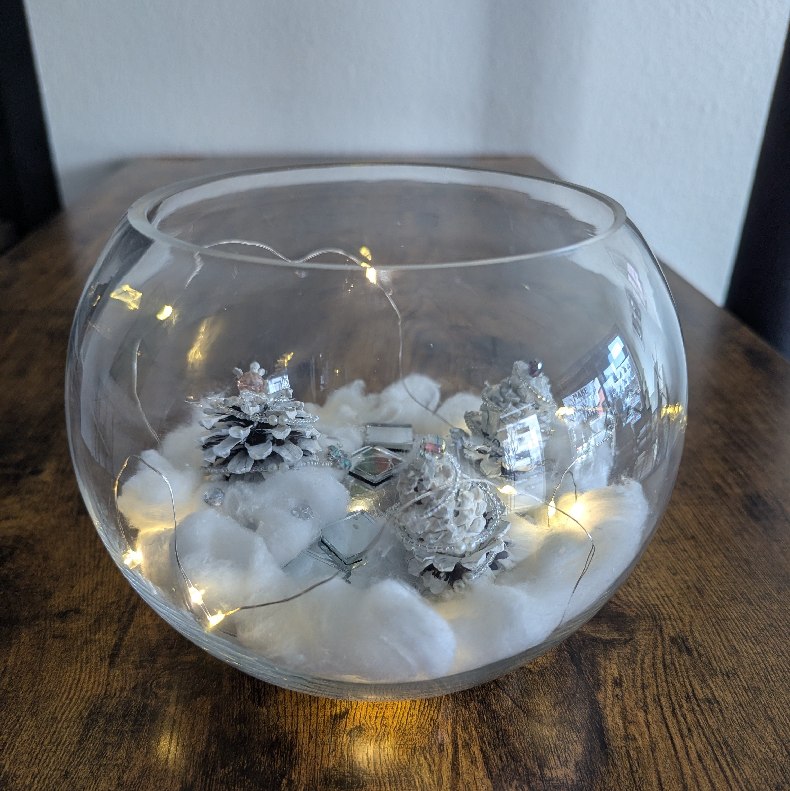 Decorative glass bowl with LED lights, snowflakes, and pinecones on a wooden surface.